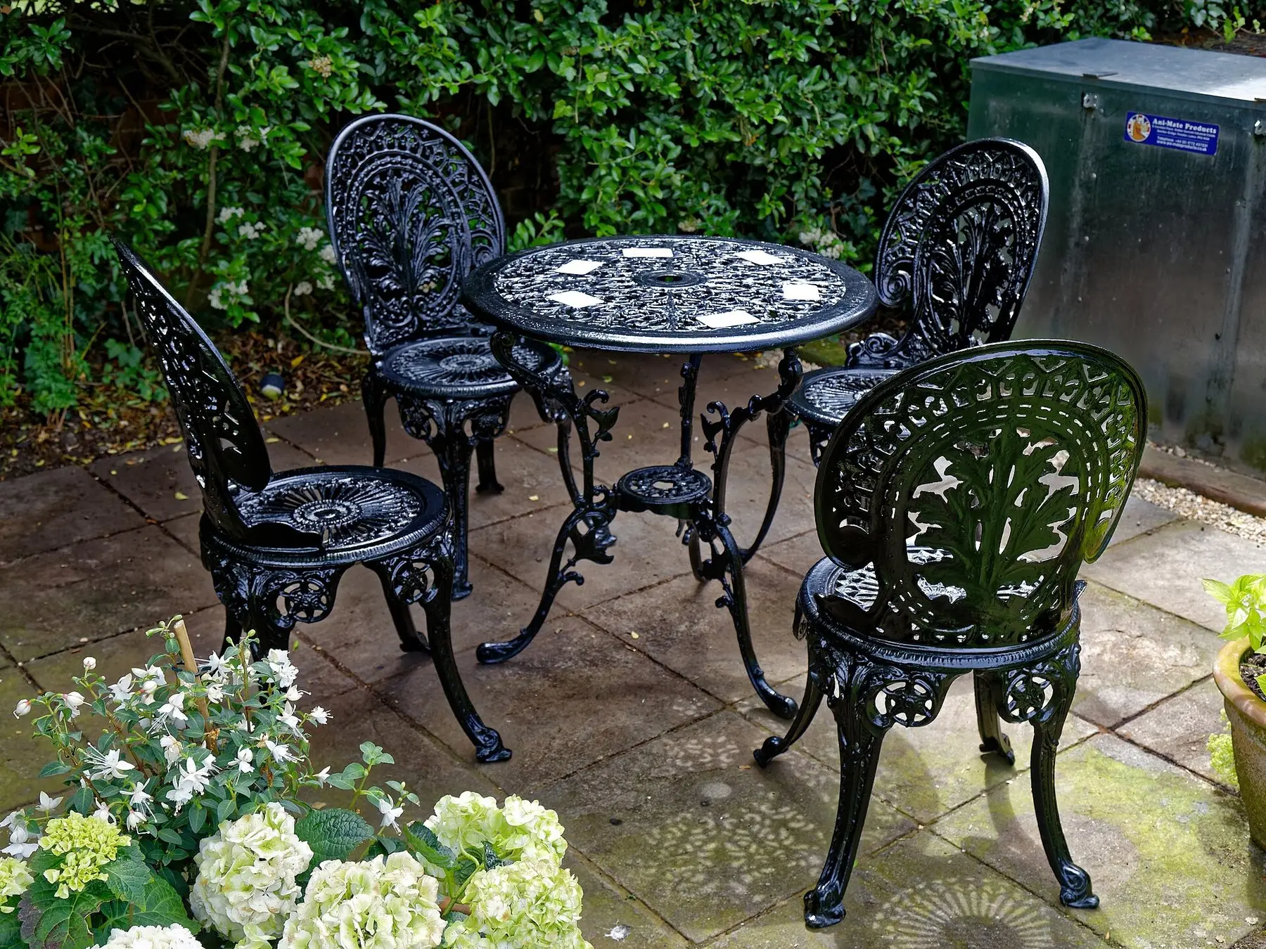 A cast iron table and chairs on a paved patio beside hydrangeas