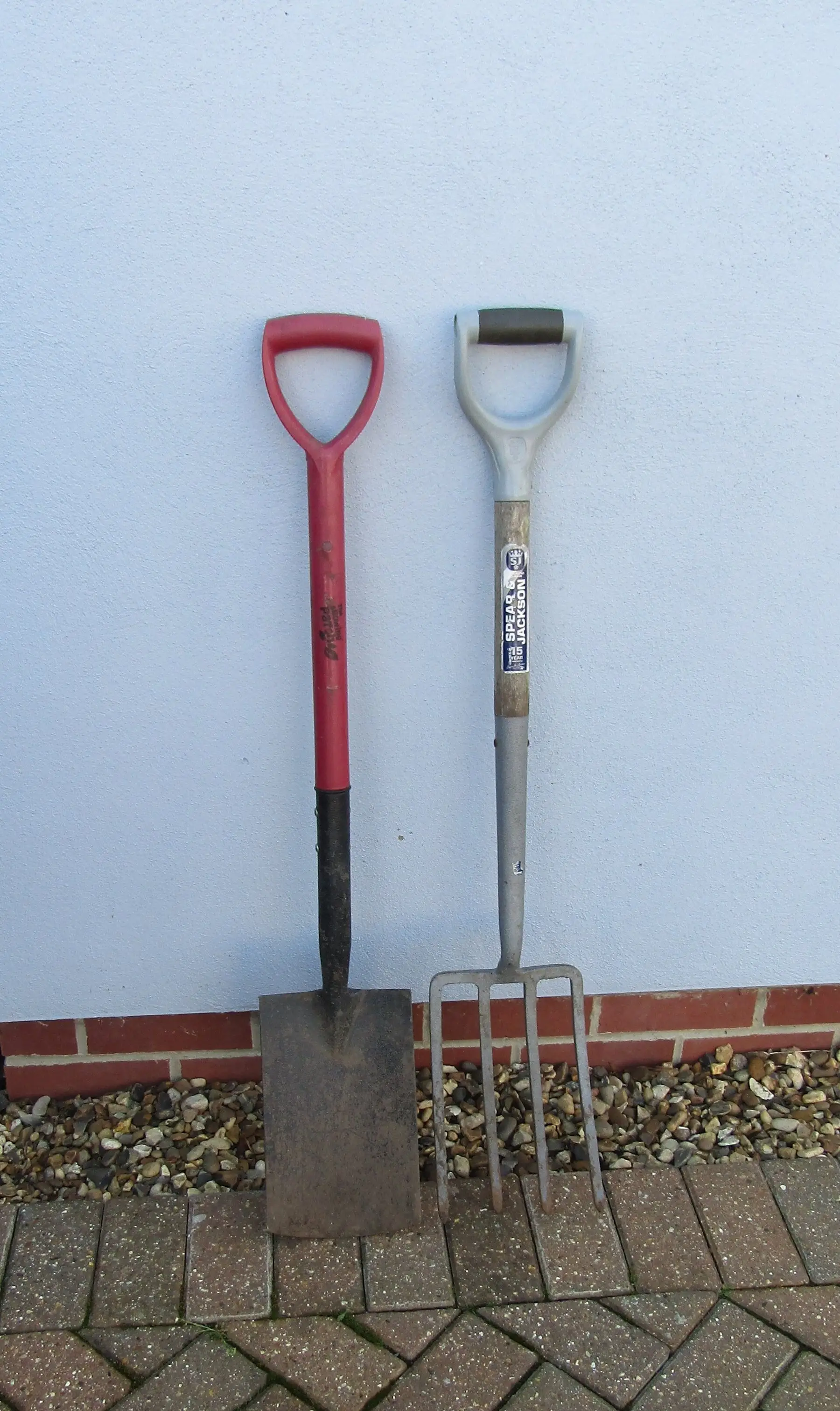 A flat-bladed garden spade standing beside a digging fork against a wall