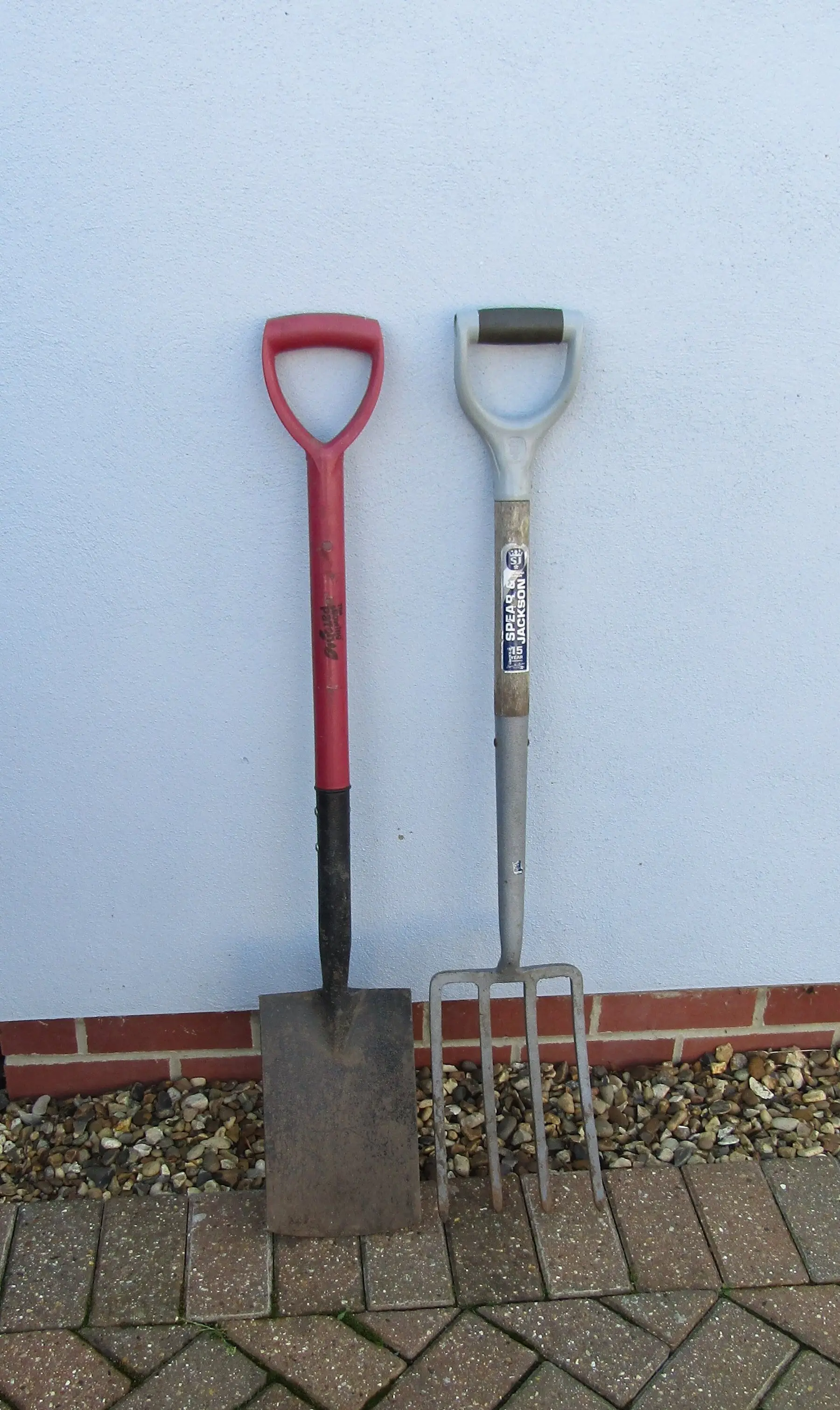 A flat-bladed garden spade standing beside a digging fork against a wall